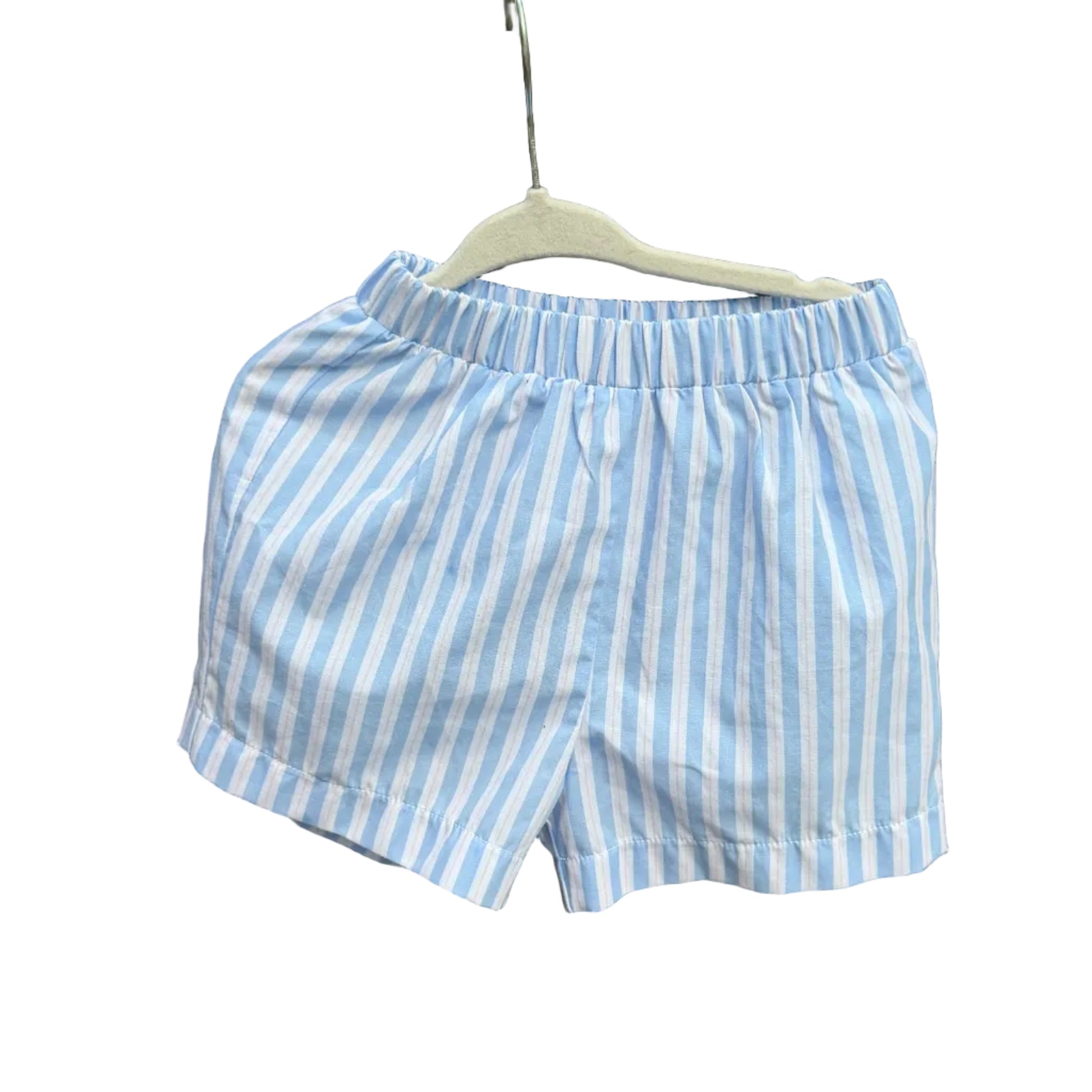 Light blue and white vertically striped boxer shorts hanging on a white plastic hanger against a rustic red-brown brick wall with green trees in the background.