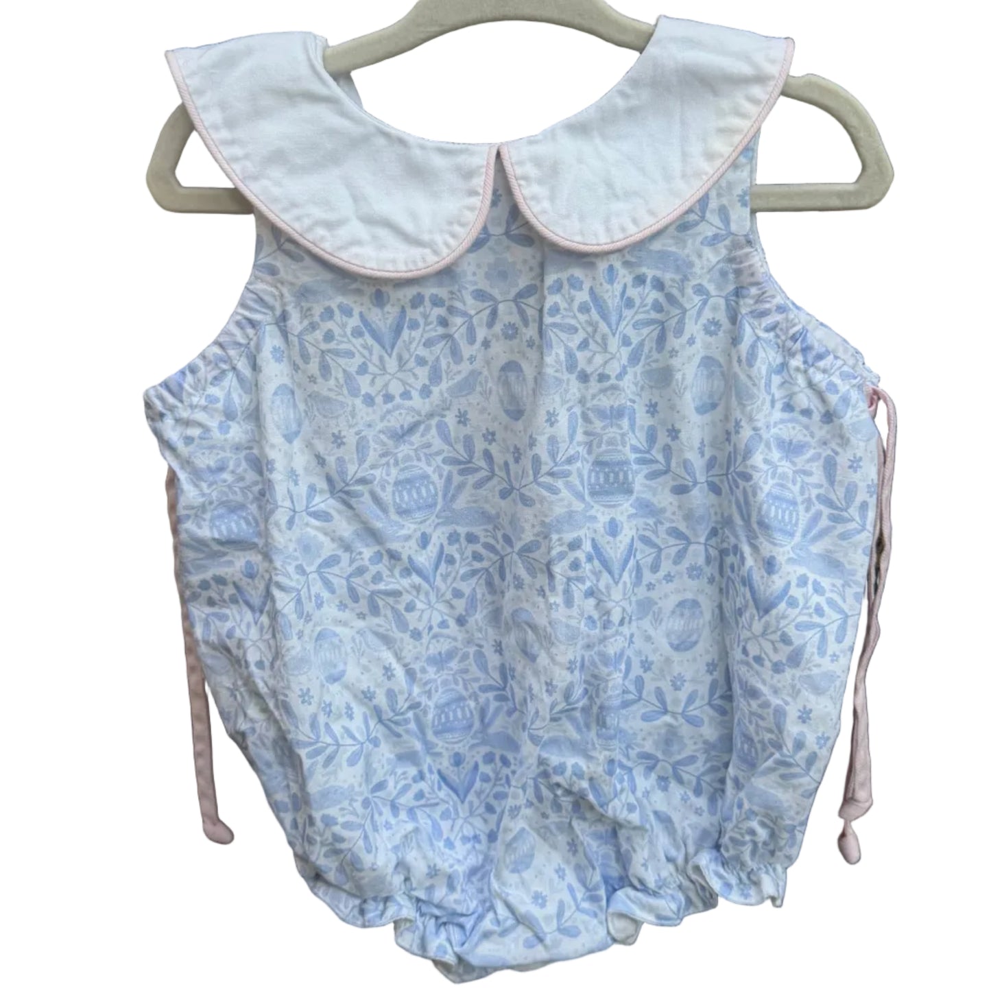 A sleeveless baby romper with a light blue and white floral pattern and a white Peter Pan collar, hanging on a white plastic hanger against a brick wall.