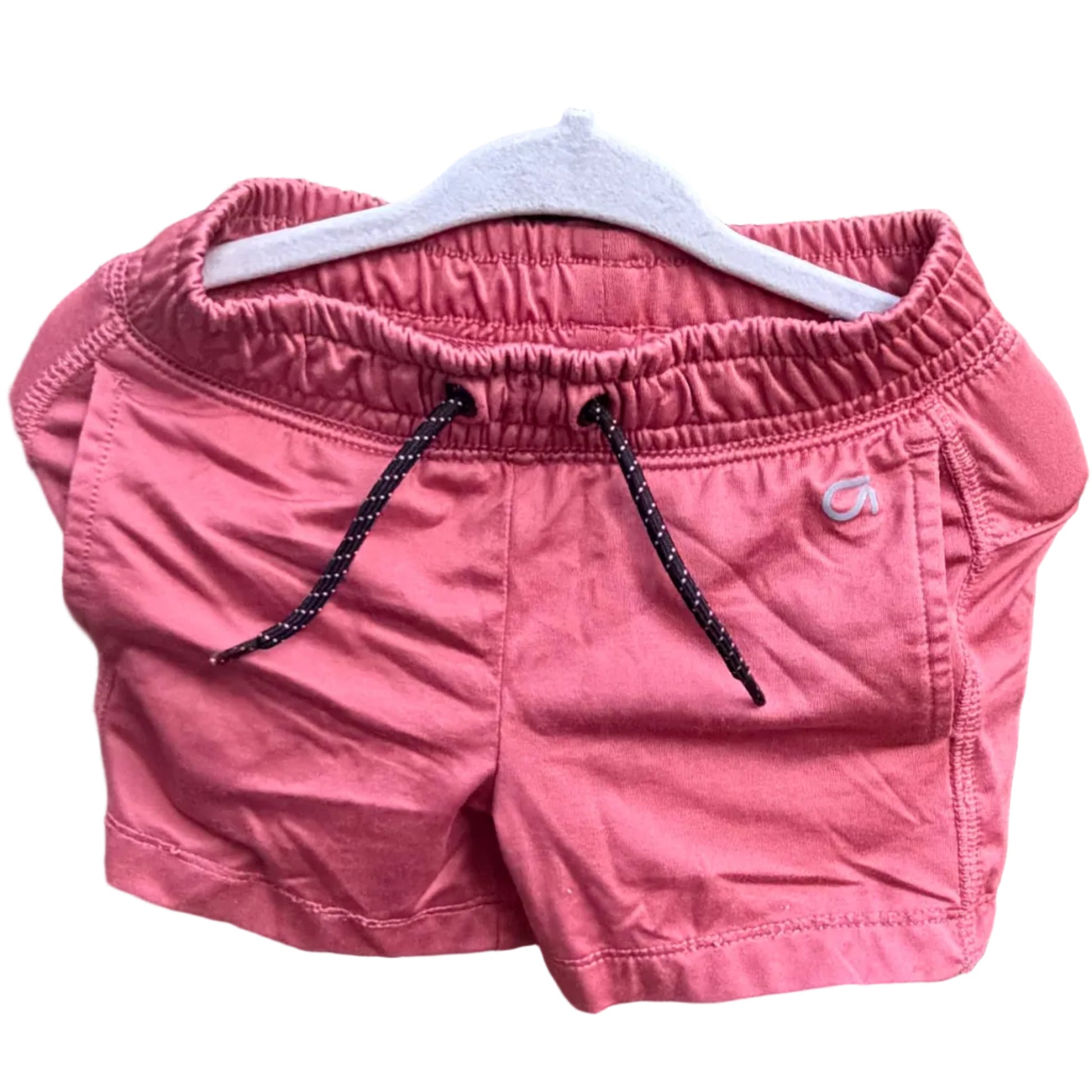Pink athletic shorts with a black drawstring, hanging on a white plastic hanger against a red-brown brick wall with green trees in the background.
