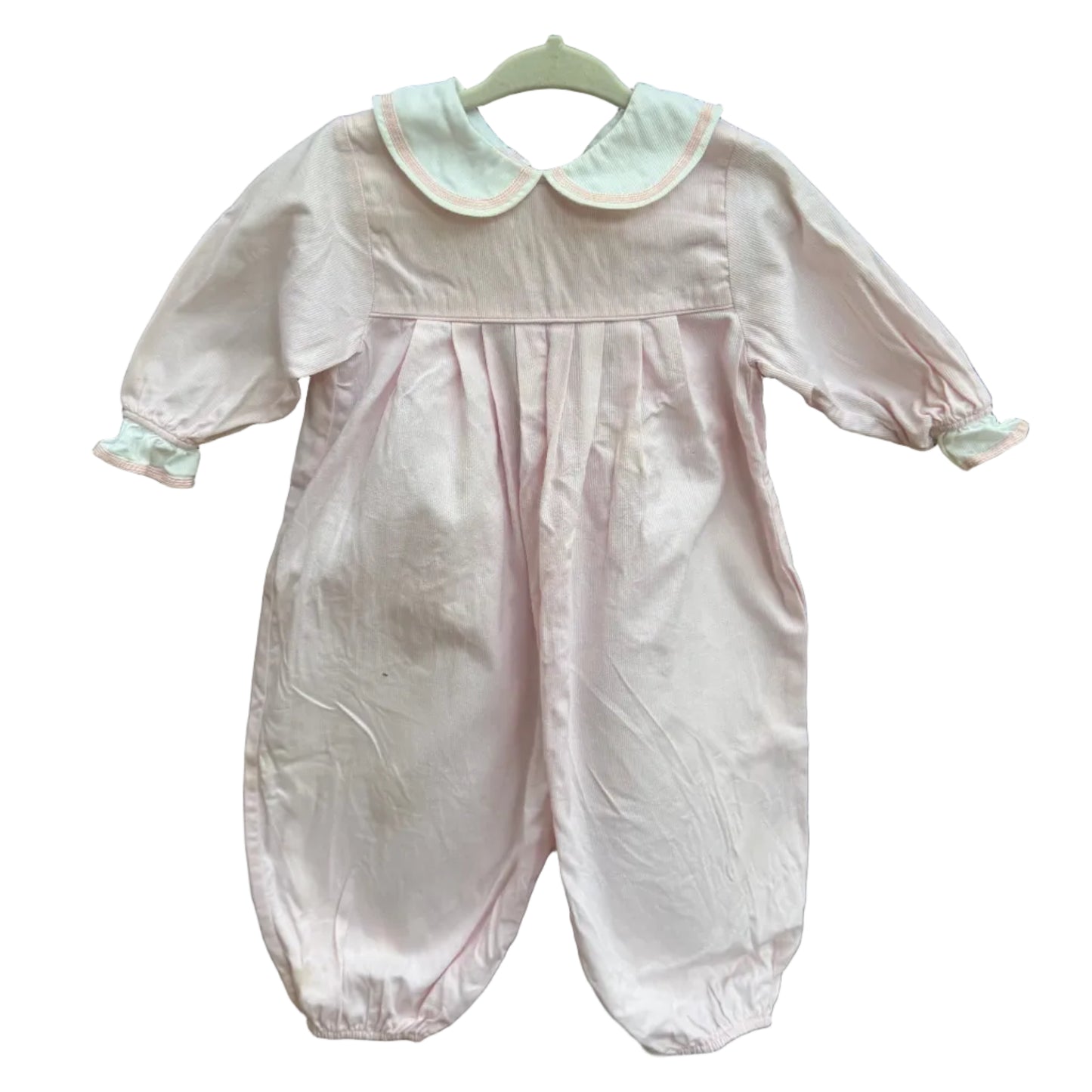 A light pink baby jumpsuit with a white Peter Pan collar and white cuffs, hanging on a white hanger against a brown brick wall with green foliage in the background.