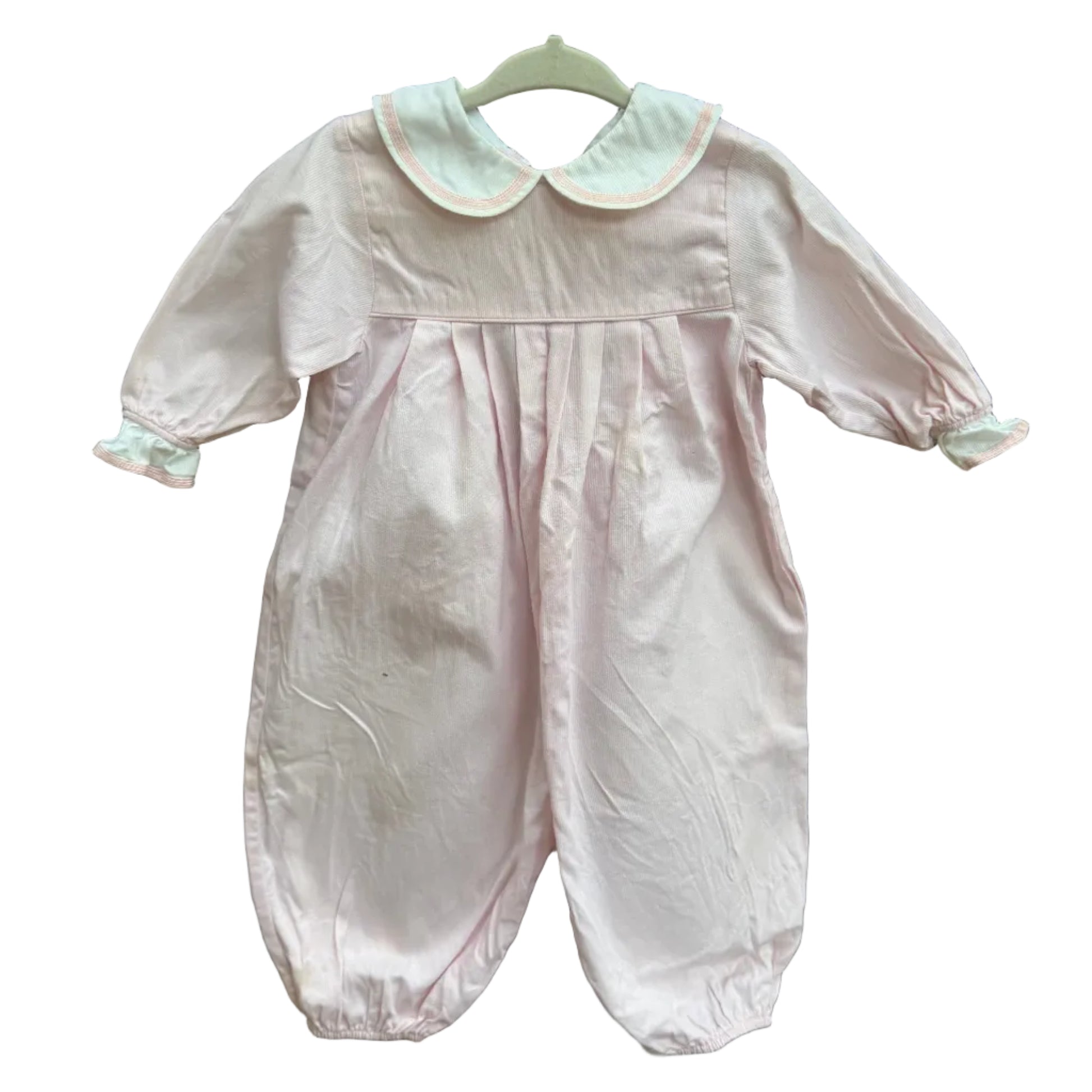 A light pink baby jumpsuit with a white Peter Pan collar and white cuffs, hanging on a white hanger against a brown brick wall with green foliage in the background.
