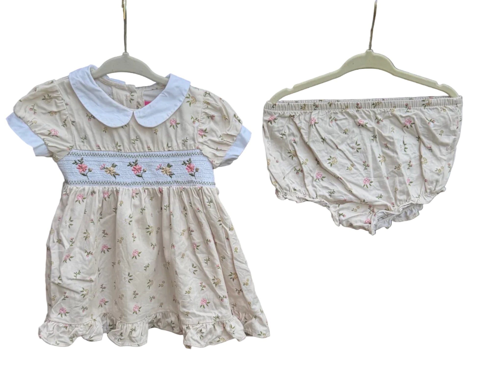 A baby girl's two-piece outfit set consisting of a cream-colored dress with a white Peter Pan collar and a matching floral bloomers, hanging on hangers against a brick wall with trees in the background.