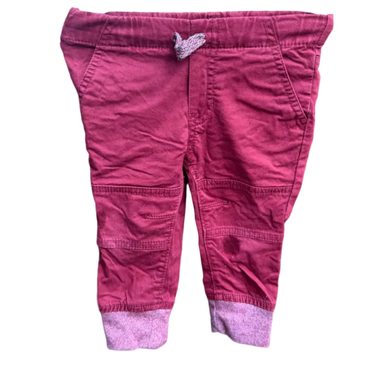 A pair of toddler's pink pants with a contrasting light pink cuff, hanging on a white plastic hanger against a brick wall.