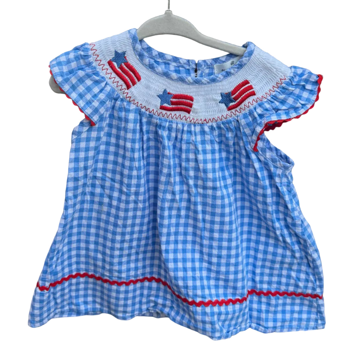 A toddler girl's blue and white gingham dress with red and white patriotic embroidery and ruffle sleeves, hanging on a white hanger against a brick wall with trees in the background.