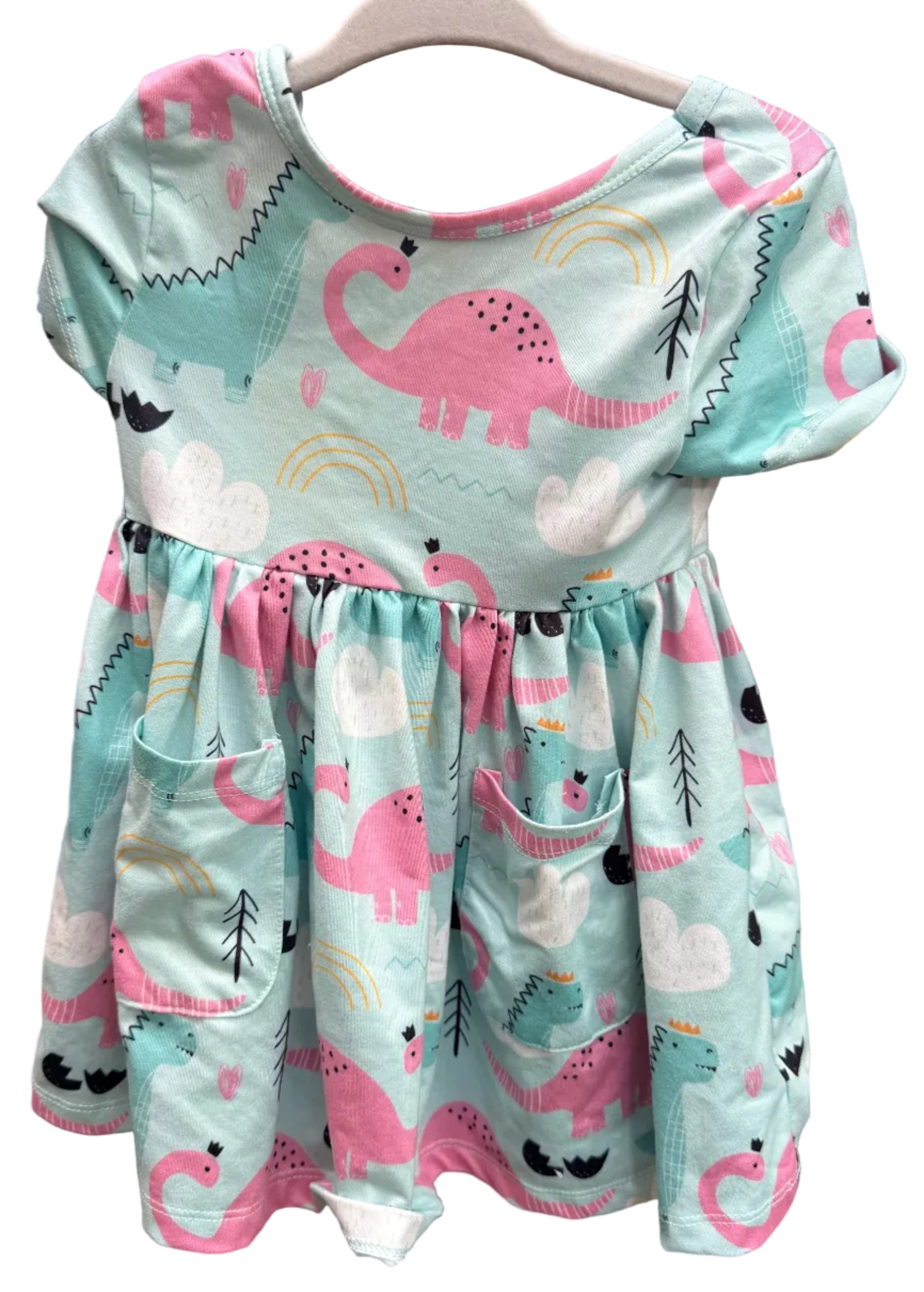 A short-sleeve children's dress with a pink dinosaur and cloud print on a mint green background, hanging on a white hanger against a brown brick wall.