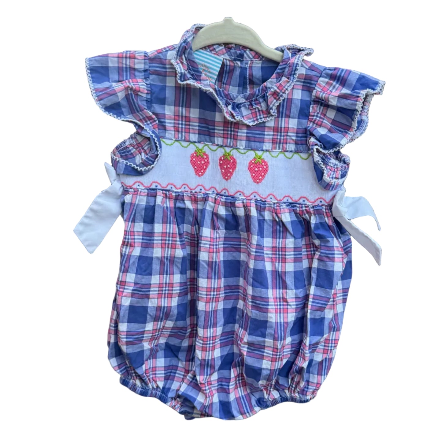 A baby romper with a blue, red, and white plaid pattern, featuring a white smocked chest panel with embroidered strawberries, hanging on a brick wall outdoors.