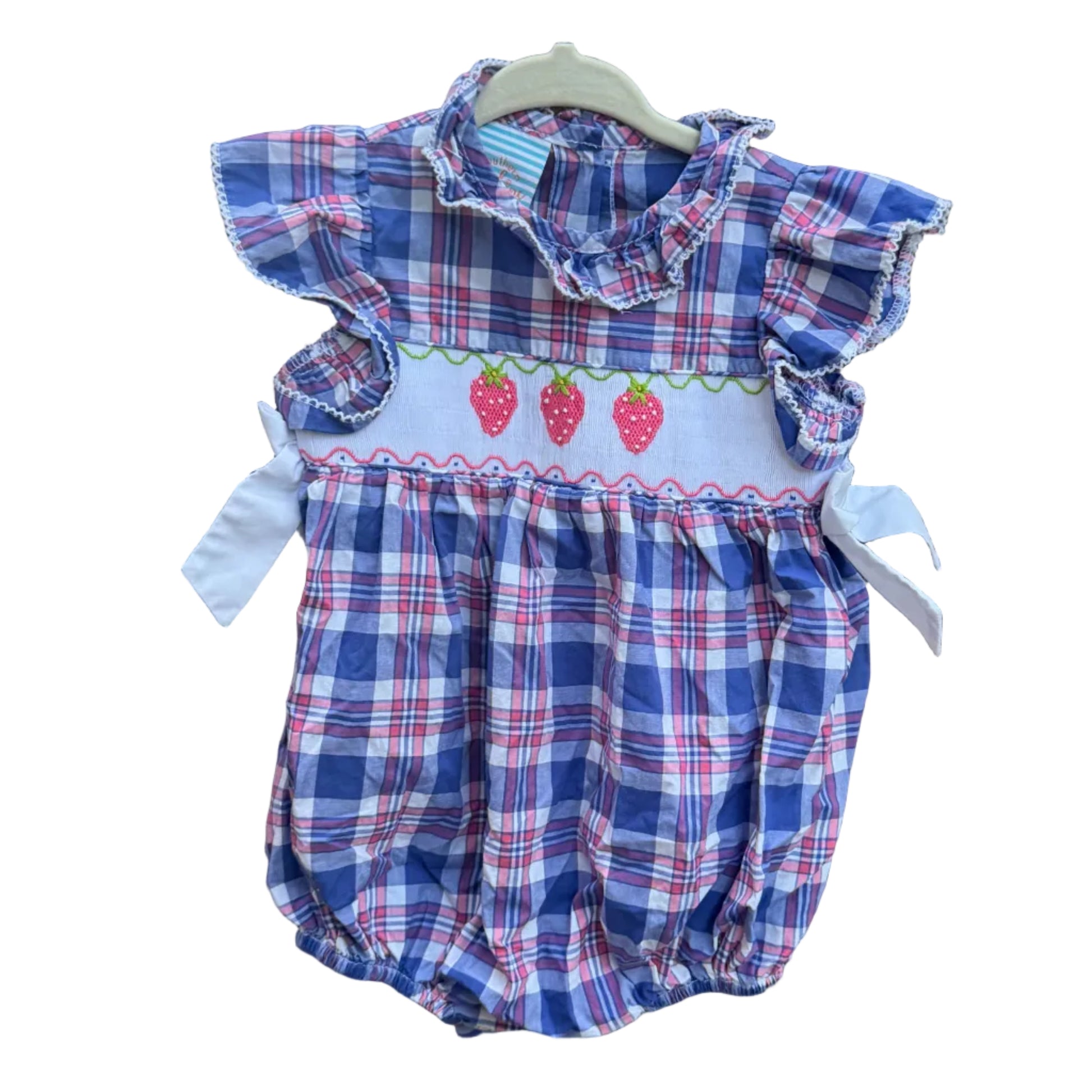 A baby romper with a blue, red, and white plaid pattern, featuring a white smocked chest panel with embroidered strawberries, hanging on a brick wall outdoors.