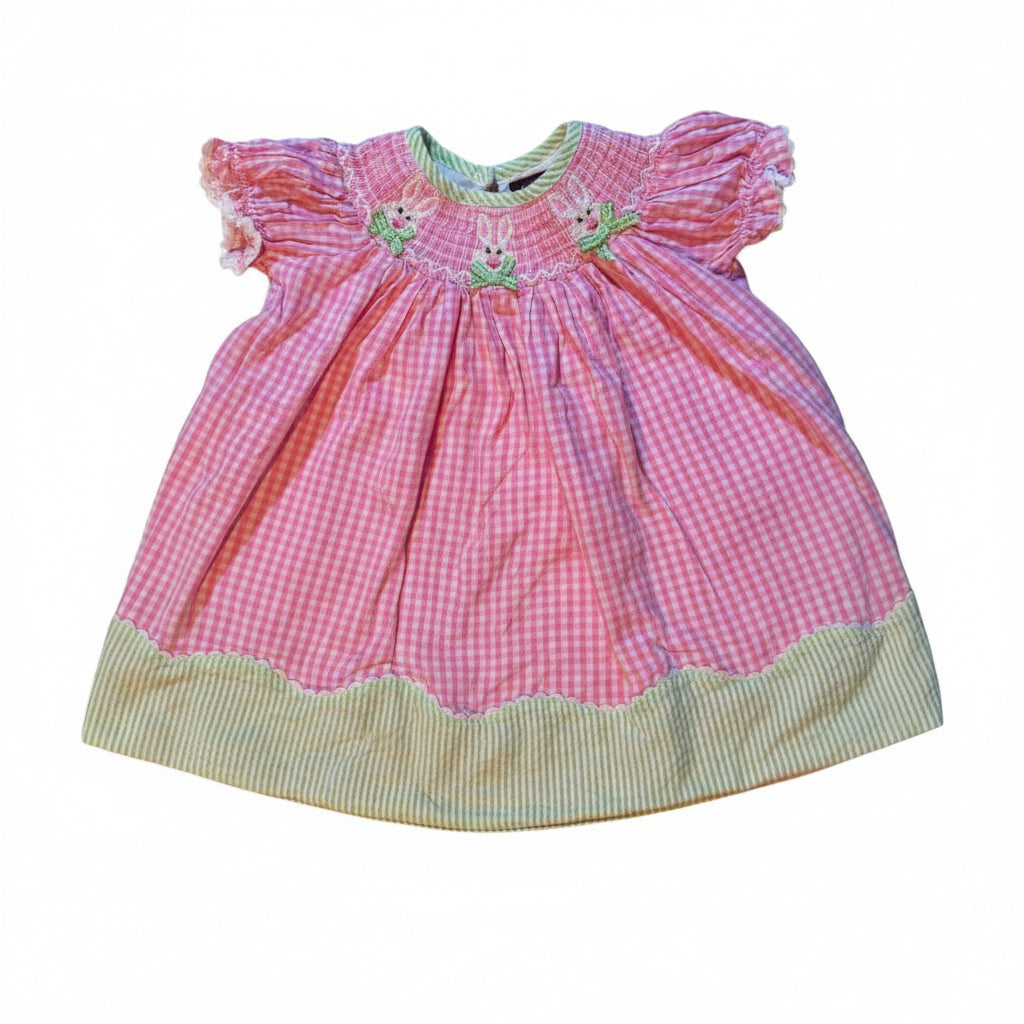 A vintage pink gingham baby bonnet with a green striped trim and embroidered floral details, laid flat on a white fabric-covered surface.