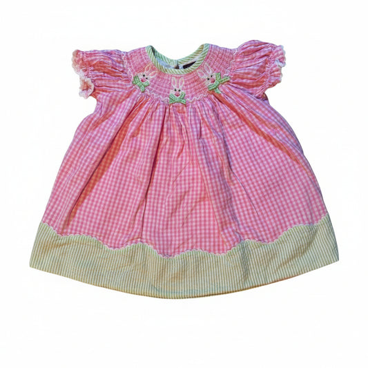 A vintage pink gingham baby bonnet with a green striped trim and embroidered floral details, laid flat on a white fabric-covered surface.