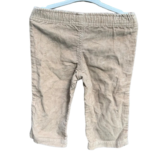 A pair of toddler's beige corduroy shorts hanging on a white plastic hanger against a red-brown brick wall with green foliage visible above.