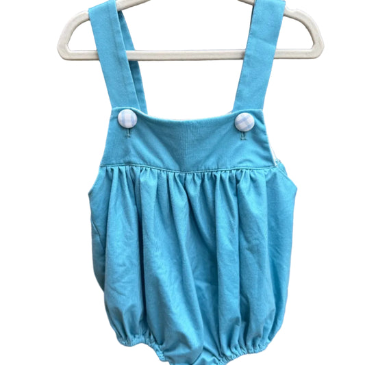 A light blue baby bubble romper with adjustable shoulder straps and white buttons, hanging on a white plastic hanger against a red-brown brick wall.