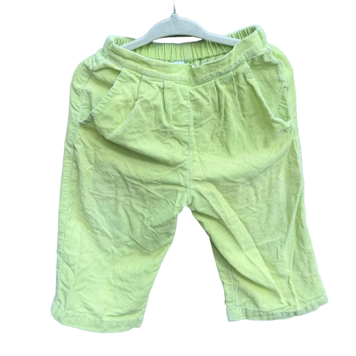 A pair of light yellow-green toddler shorts hanging on a white hanger against a red-brown brick wall with green trees in the background.