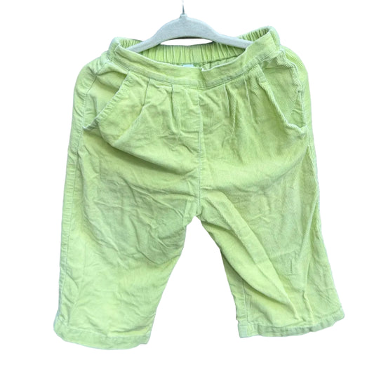 A pair of light yellow-green toddler shorts hanging on a white hanger against a red-brown brick wall with green trees in the background.
