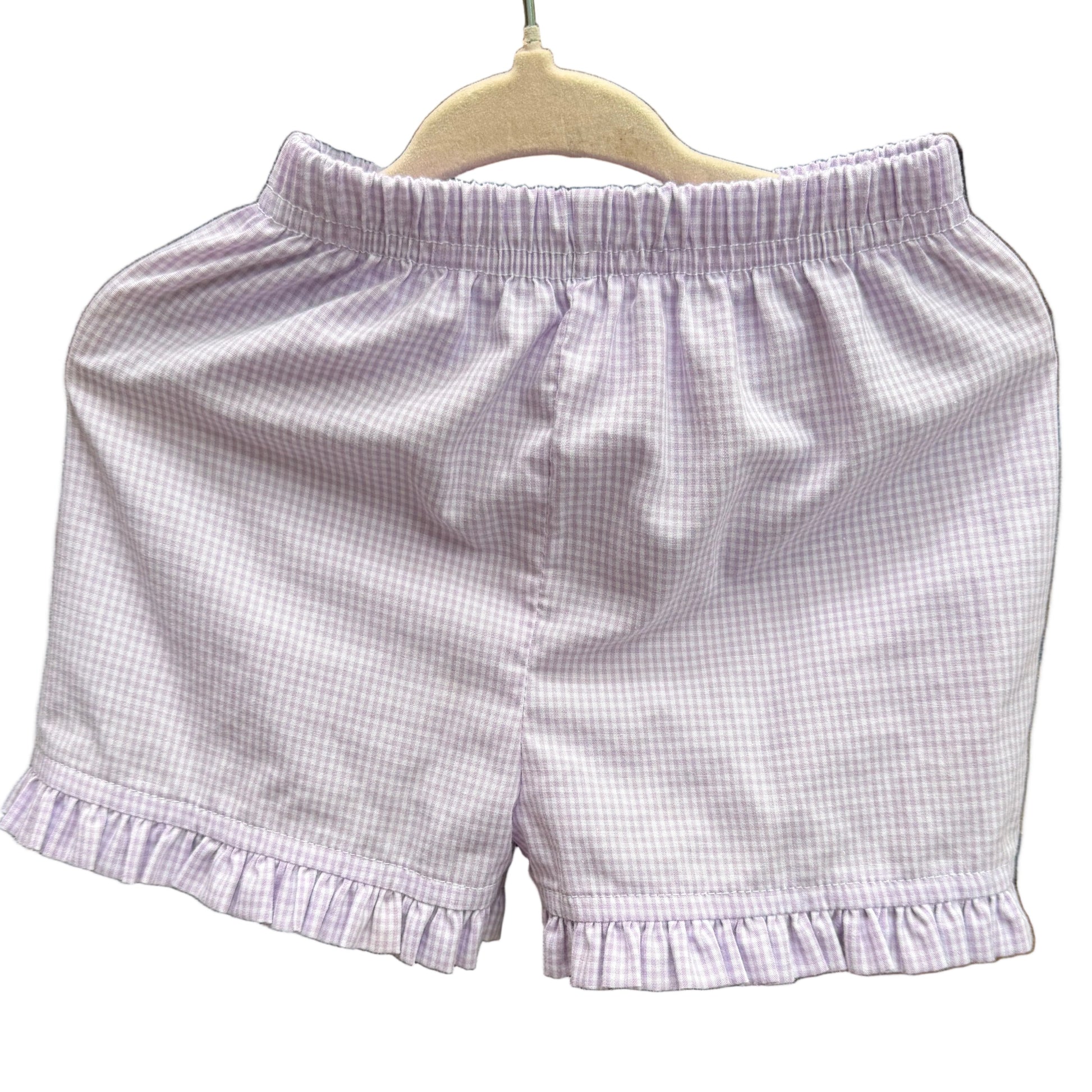 A pair of light lavender and white gingham check shorts with a ruffled hem, hanging on a wooden hanger against a yellow-brown brick wall.