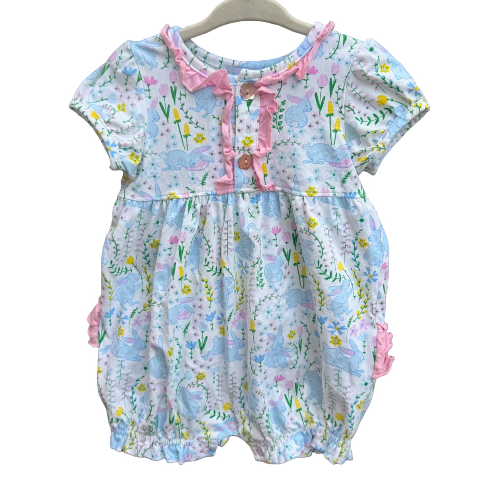 A baby romper with a light blue and white floral print, pink ruffle trim, and pink bow details, hanging on a white plastic hanger against a brick wall.