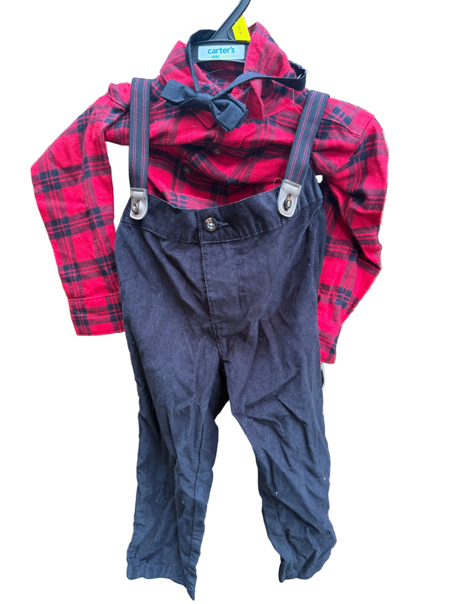 A toddler boy's clothing set consisting of a red and black plaid long-sleeve shirt, a black bow tie, and dark navy blue corduroy suspenders, hanging on a brick wall.
