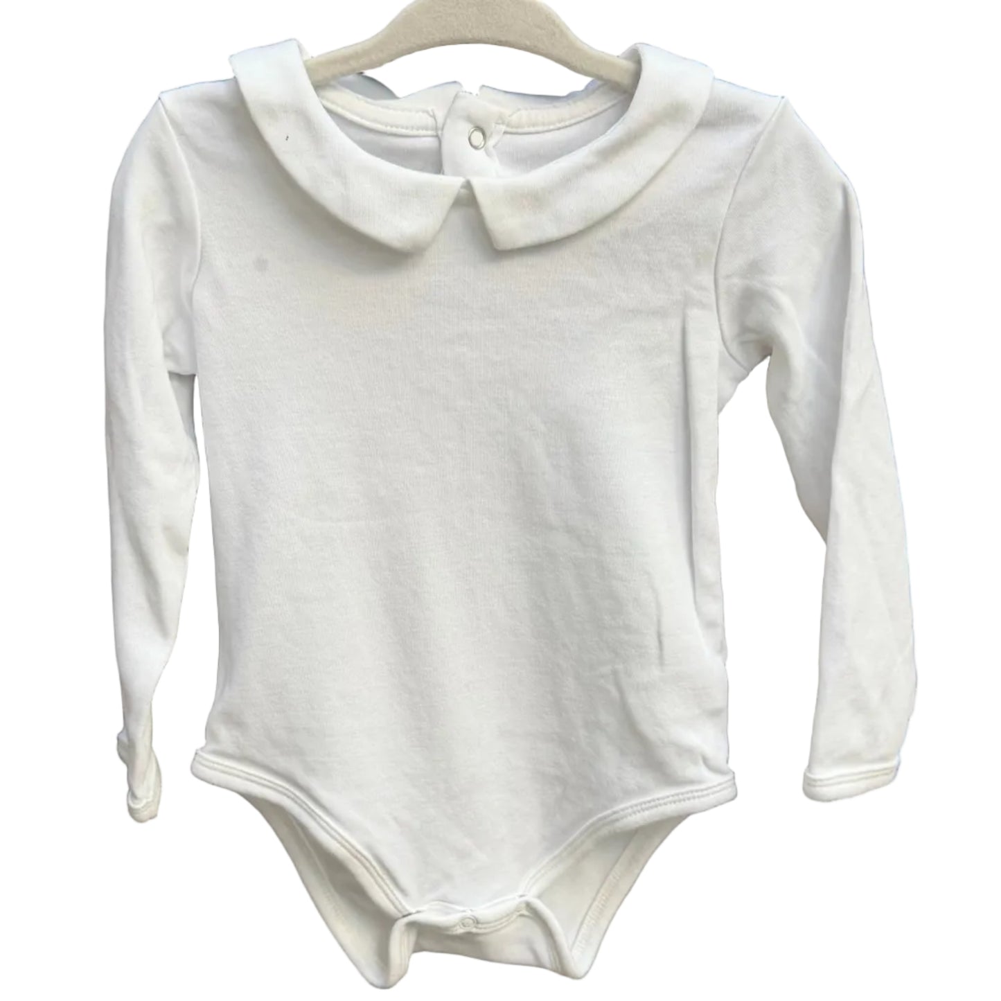 A white long-sleeve baby bodysuit with a Peter Pan collar hangs on a wooden hanger against a rustic brick wall.