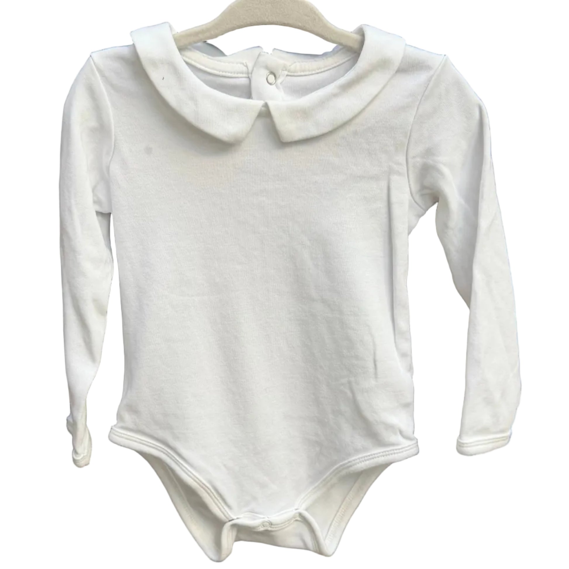 A white long-sleeve baby bodysuit with a Peter Pan collar hangs on a wooden hanger against a rustic brick wall.