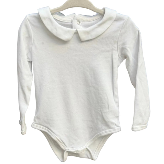 A white long-sleeve baby bodysuit with a Peter Pan collar hangs on a wooden hanger against a rustic brick wall.