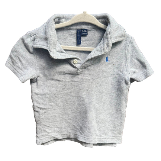 A light gray heather short-sleeve polo shirt for toddlers, featuring a small blue embroidered polo player logo on the left chest, hanging on a white plastic hanger against a red-brown brick wall.