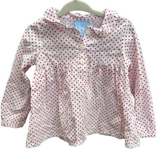 A children's long-sleeve top with a white base and small red polka dots, hanging on a white plastic hanger against a brown brick wall.