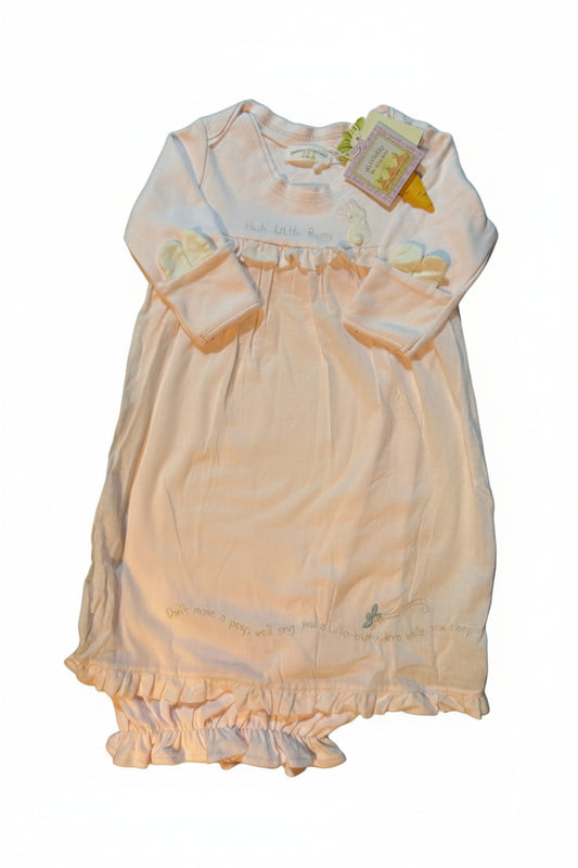 A pale pink baby romper with ruffled trim and a ruffled waistband, laid flat on a white towel. The garment has a small yellow graphic on the lower left leg and a hangtag attached.