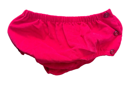 A bright red baby bloomers diaper cover with snap closures, hanging on a white plastic hanger against a brick wall background.
