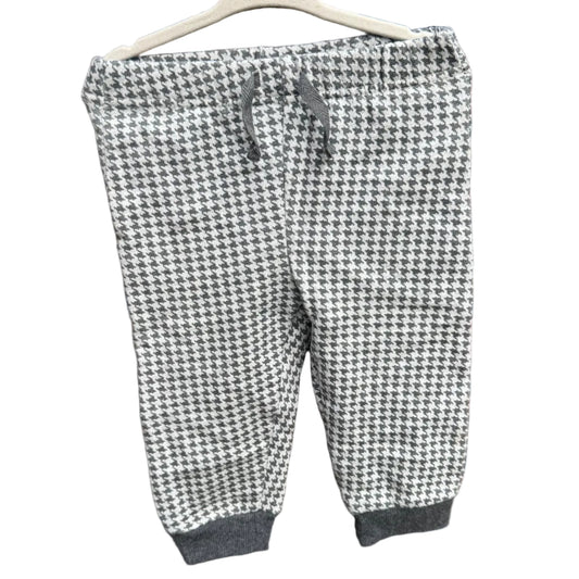 A pair of children's jogger pants with a black and white houndstooth pattern, hanging on a white plastic hanger against a red-brown brick wall.