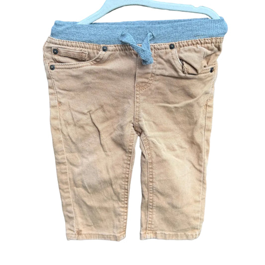 A pair of toddler boy's tan corduroy shorts with a gray elastic waistband and drawstring, hanging on a white plastic hanger against a brick wall.