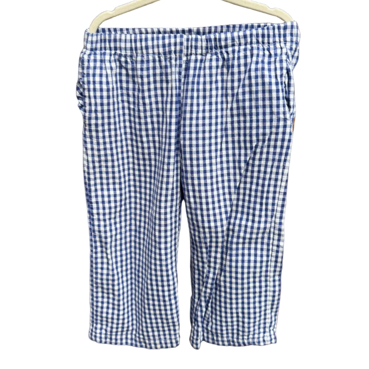 Blue and white gingham check cropped shorts hanging on a white plastic hanger against a brick wall background.