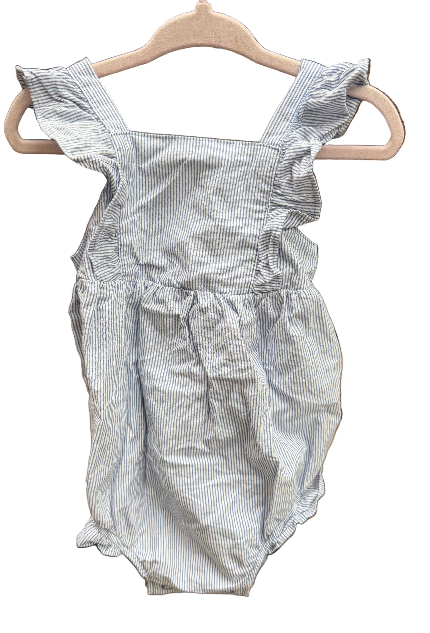 A baby romper with light blue and white vertical pinstripes, featuring ruffled shoulder straps and a smocked waist, hanging on a wooden hanger against a brick wall.