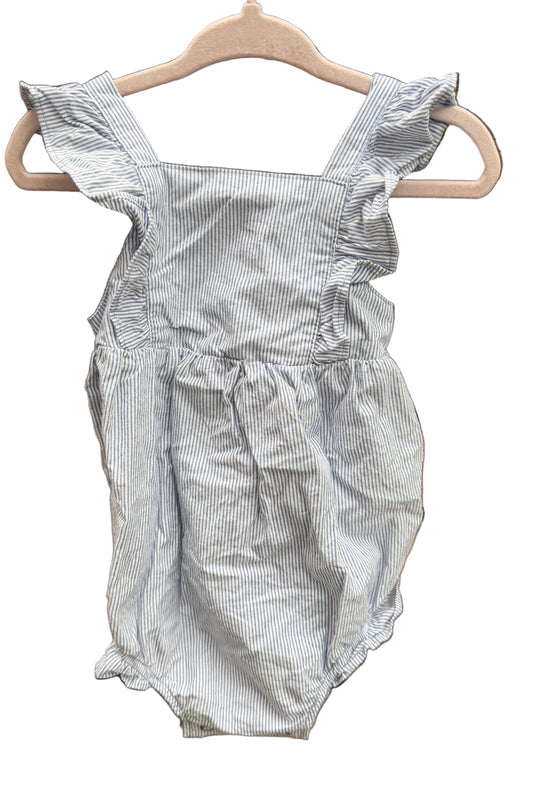A baby romper with light blue and white vertical pinstripes, featuring ruffled shoulder straps and a smocked waist, hanging on a wooden hanger against a brick wall.