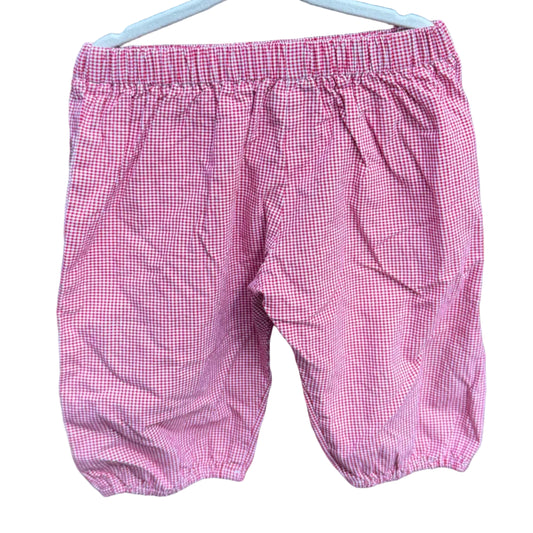 Pink and white checkered baby shorts hanging on a white plastic hanger against a brick wall.