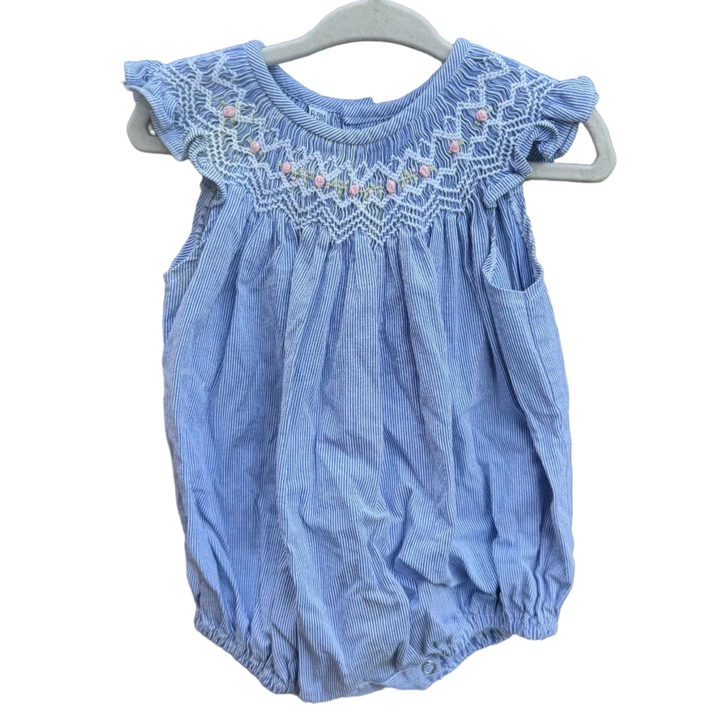 A light blue baby romper with white smocked detailing and small pink accents, hanging on a white plastic hanger against a red-brown brick wall.