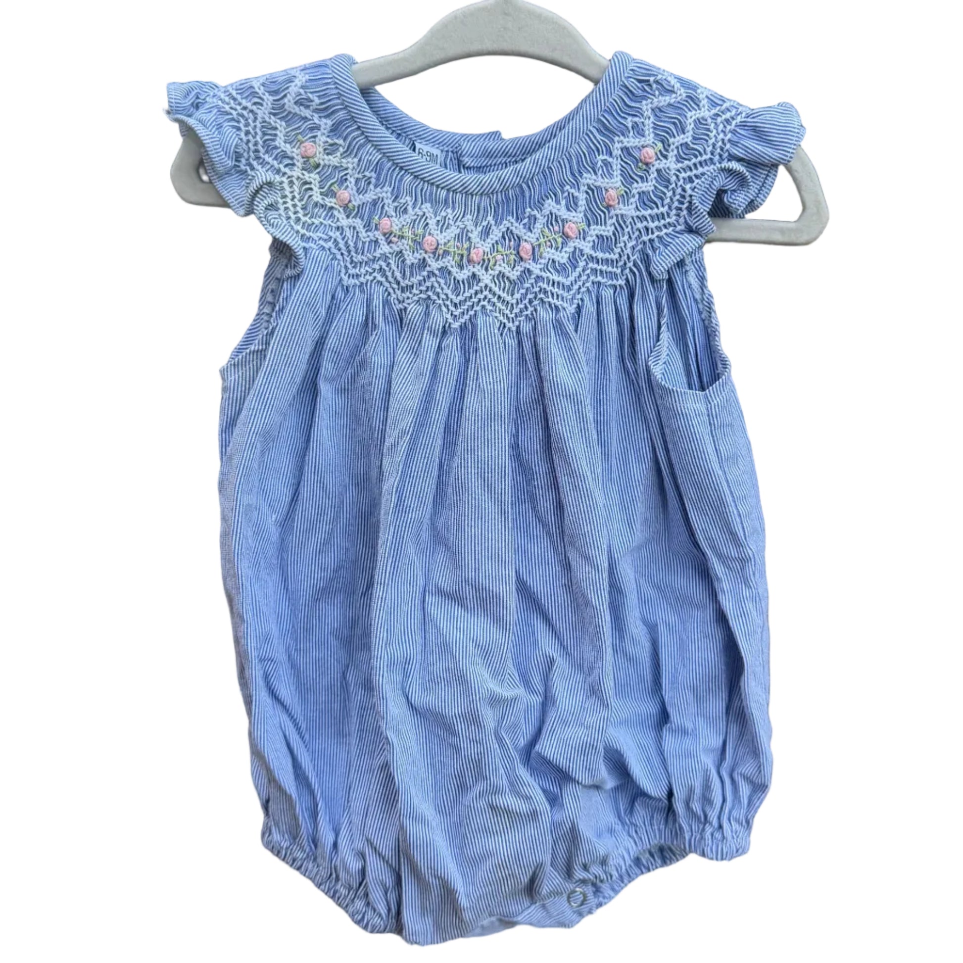 A light blue baby romper with white smocked detailing and small pink accents, hanging on a white plastic hanger against a red-brown brick wall.