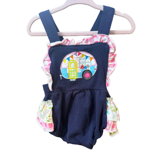 A baby romper in navy blue fabric with pink and white ruffle trim, featuring a colorful retro campervan applique on the chest, hanging on a white plastic hanger against a brick wall.