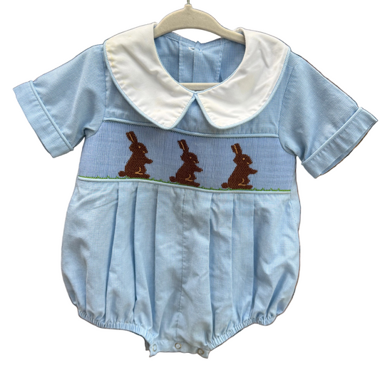 A baby short-sleeve romper in light blue denim-like fabric with a white Peter Pan collar and brown embroidered bunnies across the chest, hanging on a brick wall outdoors.