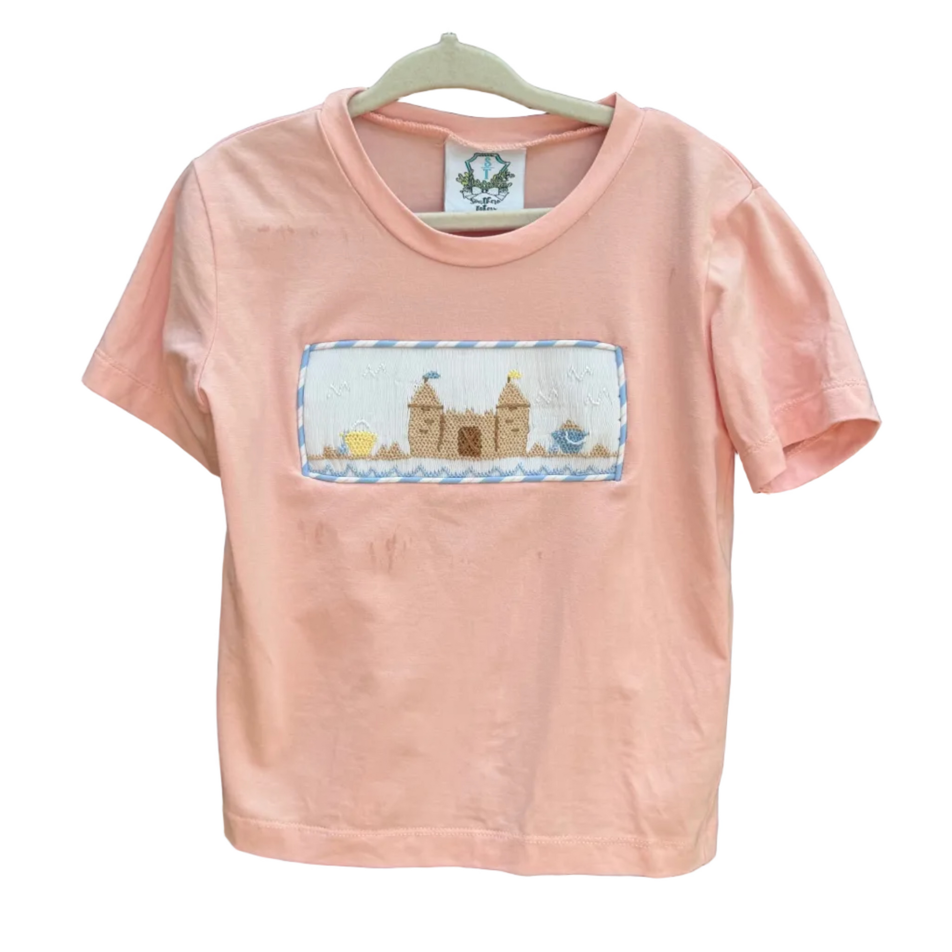 A light pink short-sleeve t-shirt with a rectangular embroidered patch depicting a church and a fountain, hanging on a white hanger against a brick wall.