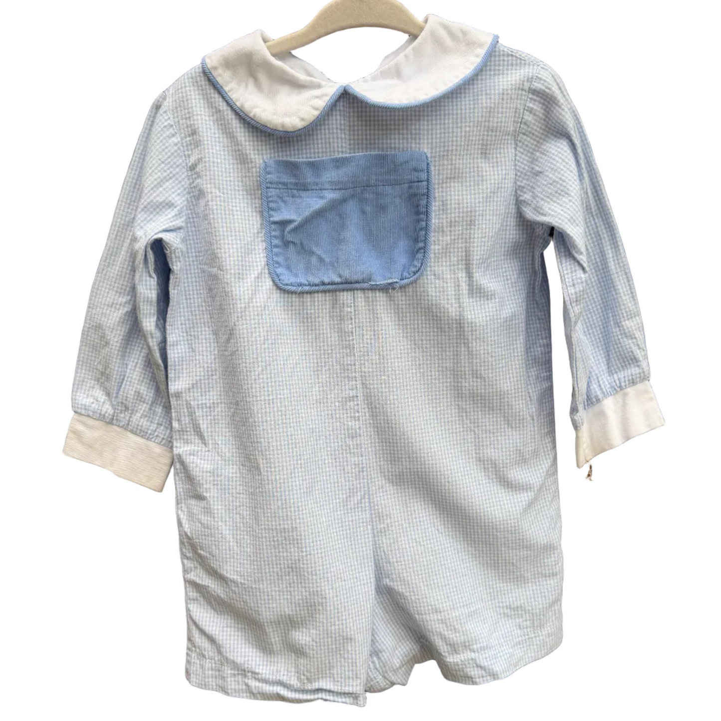 A light blue and white gingham baby romper with a solid light blue chest pocket and white collar, hanging on a wooden hanger against a brick wall.
