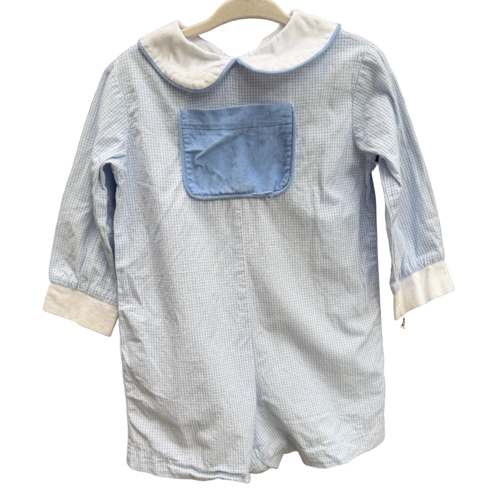 A light blue and white gingham baby romper with a solid light blue chest pocket and white collar, hanging on a wooden hanger against a brick wall.