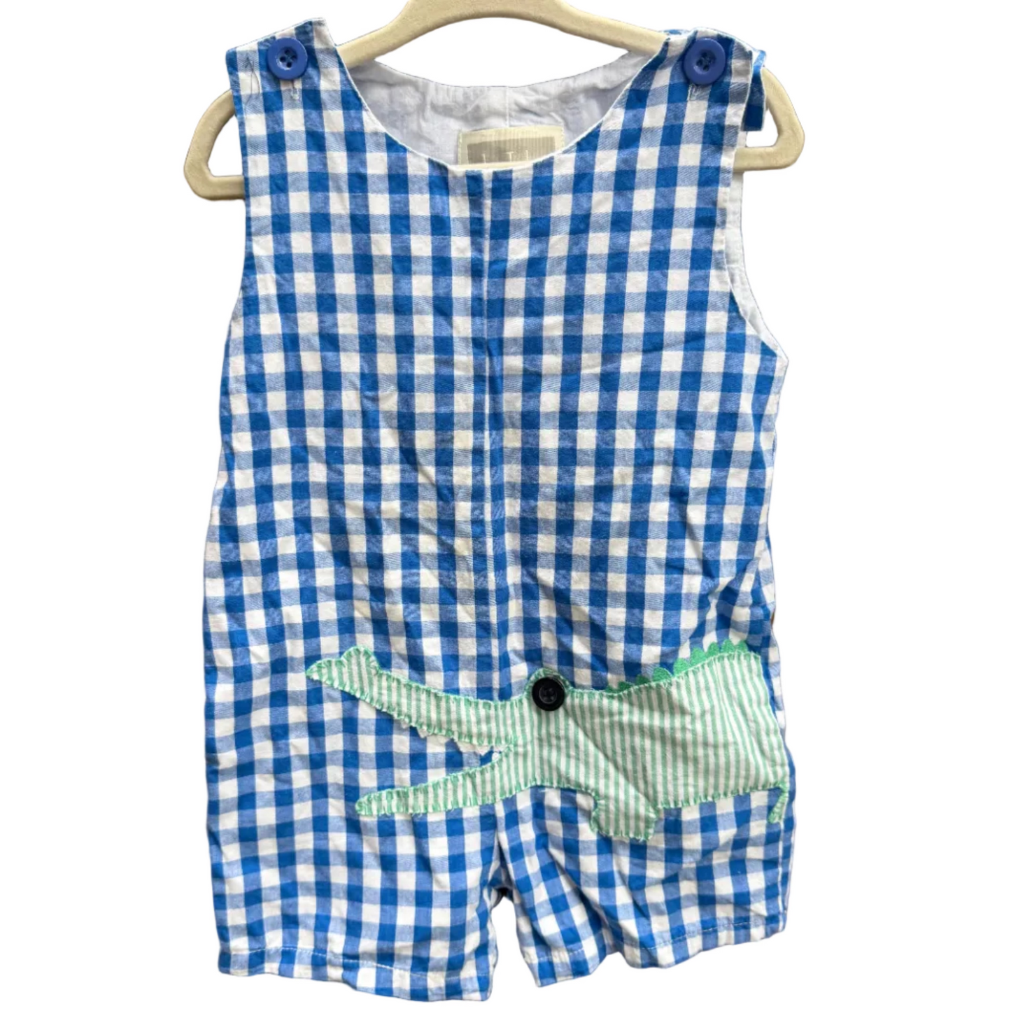 A sleeveless baby romper with a blue and white gingham pattern, featuring a green and white striped patchwork front panel with a black button, hanging on a wooden hanger against a brick wall.