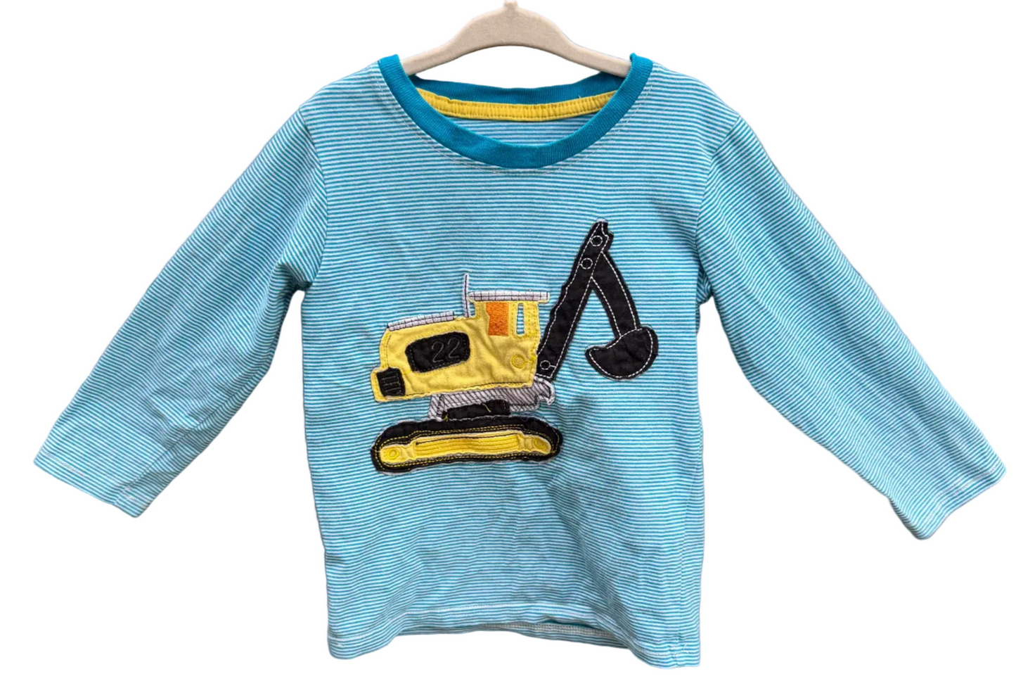 A light blue long-sleeve children's shirt with a yellow and black excavator graphic on the front, hanging on a wooden hanger against a brick wall.
