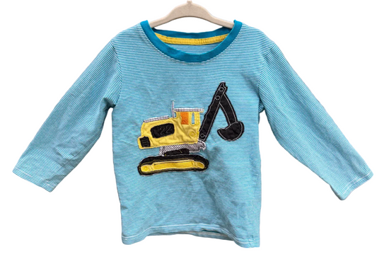 A light blue long-sleeve children's shirt with a yellow and black excavator graphic on the front, hanging on a wooden hanger against a brick wall.