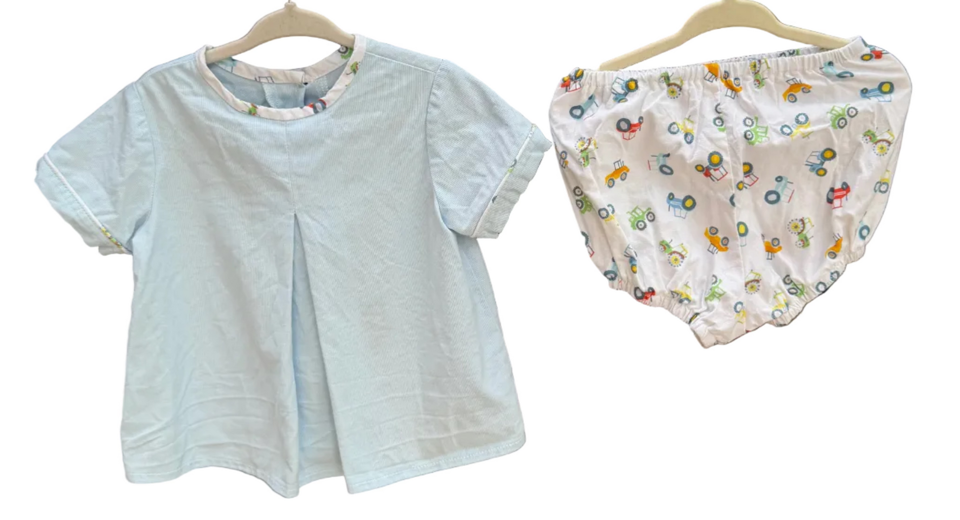 A baby outfit set consisting of a white short-sleeve top and white patterned bloomers, hanging on wooden hangers against a brick wall with trees in the background.