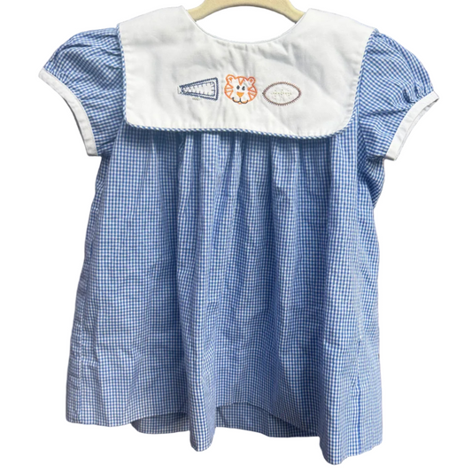 A blue and white gingham baby dress with a white bib collar featuring embroidered initials and a small cat patch, hanging on a wooden hanger against a brick wall.
