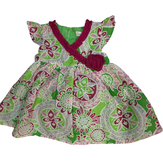 A toddler girl's sleeveless dress with a green, pink, and white floral paisley pattern, featuring a magenta pink crocheted V-neck trim and a matching flower applique on the chest.